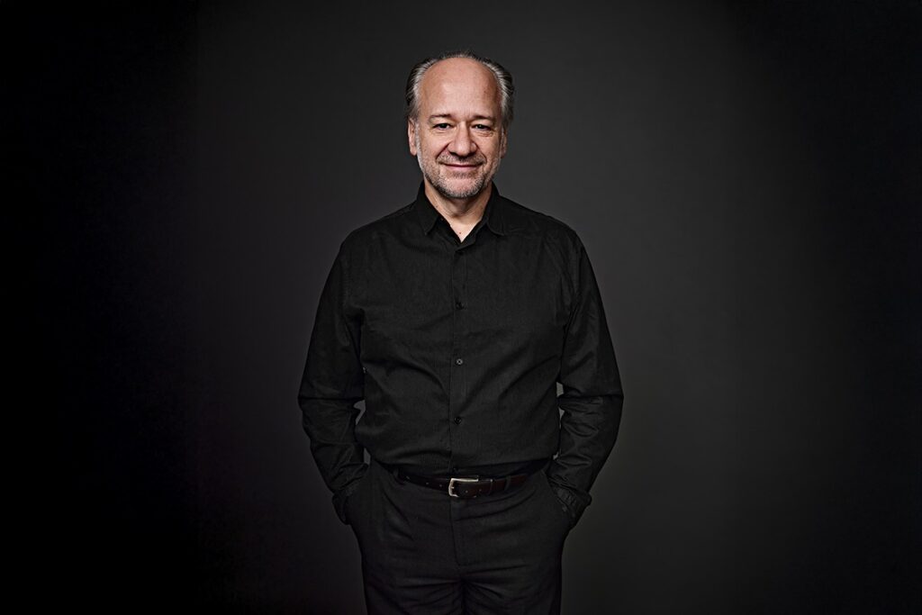 Um homem de meia-idade, com cabelos grisalhos curtos e pele clara, está em pé contra um fundo escuro. Ele está usando uma camisa preta de botões e calças pretas, com as mãos nos bolsos. Ele tem um leve sorriso no rosto e está olhando diretamente para a câmera. A iluminação é suave, iluminando o rosto do homem e criando um leve brilho em seus cabelos. O fundo escuro e a iluminação dramática criam um clima formal e profissional.