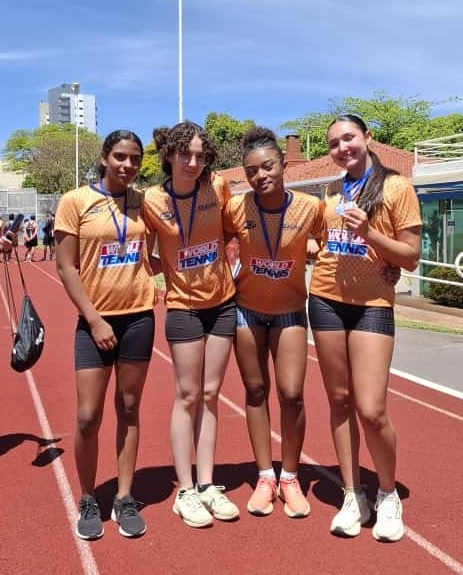 A imagem mostra quatro jovens atletas em uma pista de atletismo, vestindo uniformes laranja com o logo "WORLD TENNIS". Elas posam para a foto, sorrindo e mostrando medalhas. As atletas têm diferentes tons de pele e cabelos, sugerindo diversidade. O fundo revela uma área externa com céu azul, árvores e edifícios ao longe, indicando um dia ensolarado e um evento esportivo.
