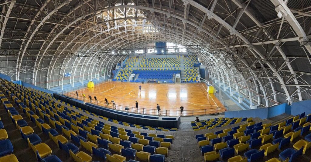 A imagem mostra o interior de uma quadra esportiva, provavelmente de basquete, com arquibancadas em formato de U.As arquibancadas são compostas por assentos nas cores azul e amarelo, dispostos em vários níveis. O piso da quadra é de madeira clara e polida, com as marcações típicas de uma quadra de basquete. Há algumas pessoas na quadra, possivelmente jogadores ou pessoal de manutenção.O teto da quadra é uma estrutura complexa de metal e madeira, com arcos que se estendem por toda a extensão do espaço, criando um visual industrial e robusto. A iluminação parece ser artificial, com lâmpadas distribuídas pela estrutura do teto. Ao fundo, é possível ver mais arquibancadas e parte da estrutura externa do ginásio.