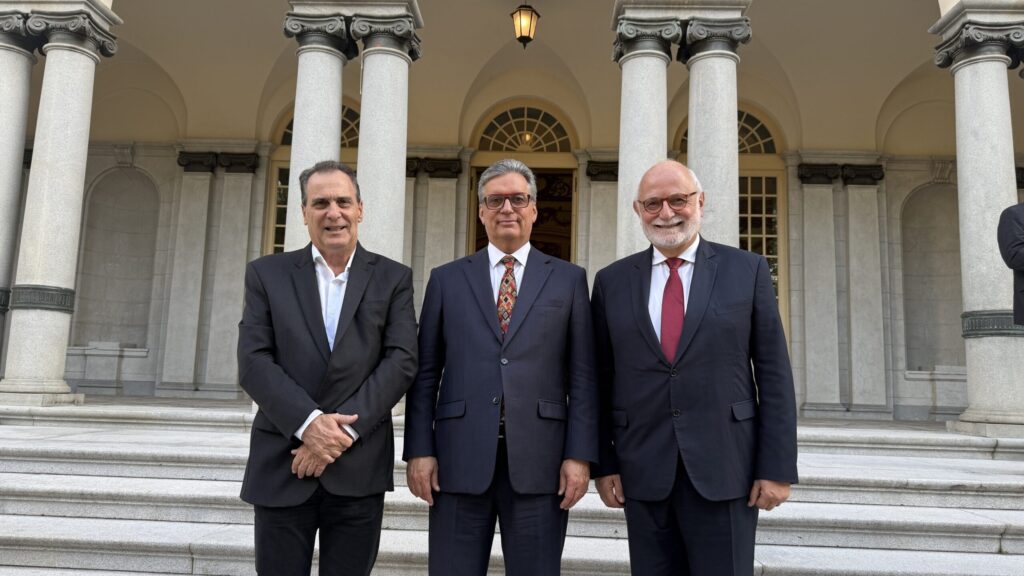 A imagem mostra três homens em frente a um edifício com colunas. Todos os homens estão vestidos com ternos escuros, e todos eles estão sorrindo. O homem da esquerda está usando uma camisa branca, enquanto os outros dois homens estão usando camisas brancas com gravatas. O homem do meio está usando um terno azul escuro com uma gravata estampada. O homem da direita está usando um terno azul escuro com uma gravata vermelha. O edifício atrás deles tem uma entrada arqueada com uma janela acima dela. Há também um candeeiro pendurado acima da entrada.