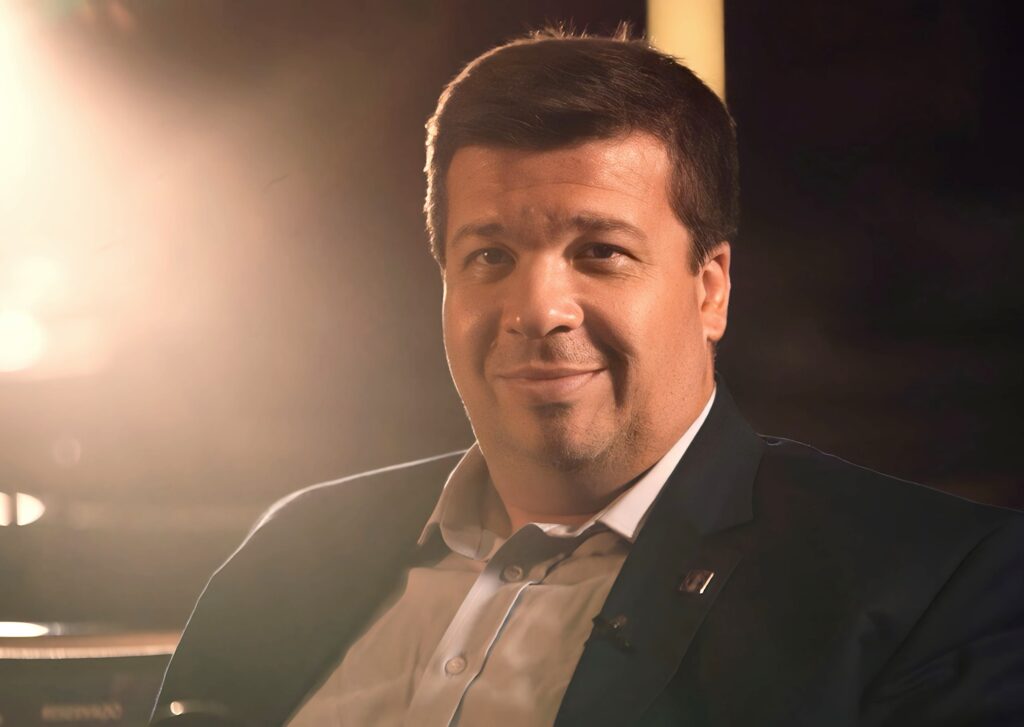 A imagem mostra um close-up de um homem sorrindo para a câmera. Ele está vestindo um terno escuro sobre uma camisa clara. A iluminação é dramática, com uma luz forte vindo da esquerda que realça seu rosto e cria um brilho suave em seu cabelo. O fundo está escuro e desfocado, com alguns pontos de luz que sugerem um ambiente interno. Há um pequeno broche preso à lapela do terno dele.