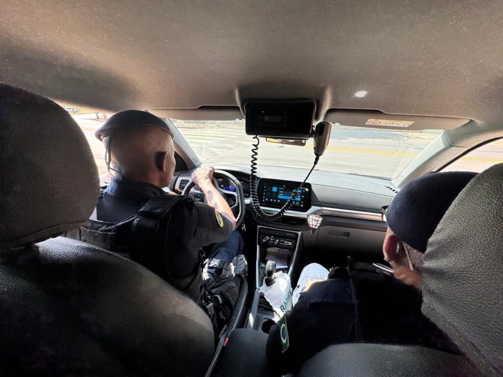 A imagem mostra o interior de um veículo, possivelmente uma viatura policial, vista a partir do banco traseiro. No banco da frente, há dois homens vestidos com uniformes escuros e boinas, que parecem ser policiais. O motorista está segurando o volante com as duas mãos e olhando para a frente, concentrado na direção. Ao lado dele, outro policial está parcialmente visível, também de uniforme e boina. O painel do carro é moderno, com telas digitais e um rádio comunicador fixado acima do painel, com um cabo espiralado. A luz do dia entra pela frente do veículo, iluminando parcialmente o interior. Ao fundo, pela janela dianteira, é possível ver uma rua com algumas construções. A imagem transmite uma sensação de ação e vigilância, típica de uma patrulha policial em andamento.