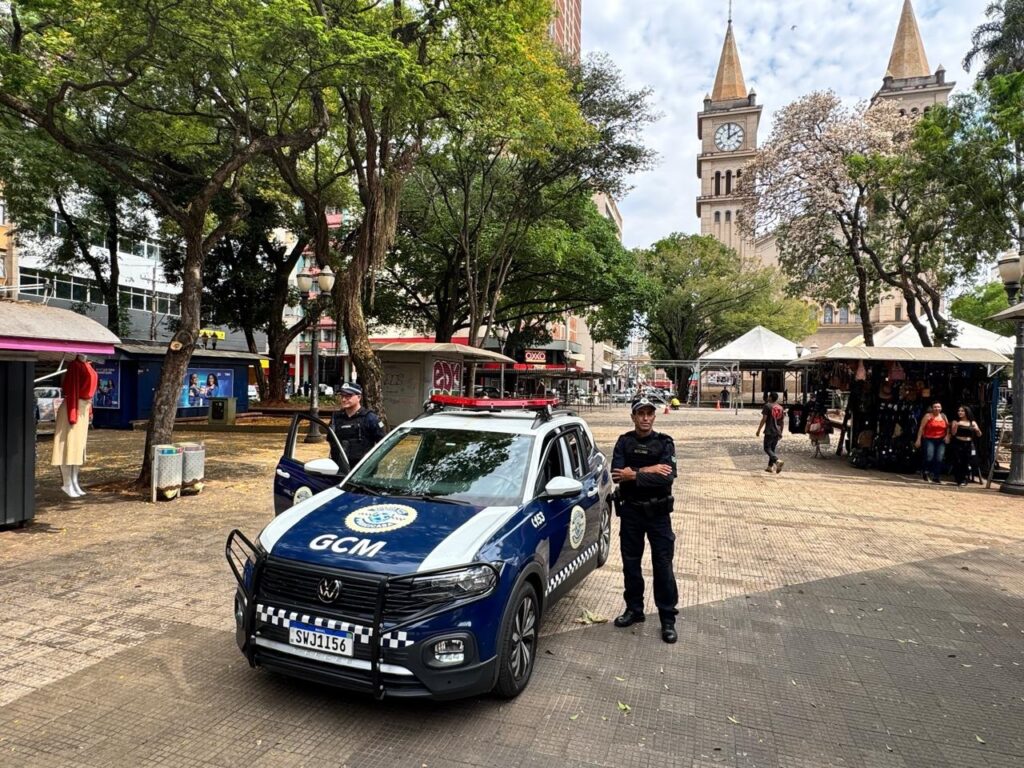A imagem mostra dois guardas municipais em frente a uma viatura da Guarda Civil Municipal (GCM). A viatura é um Volkswagen T-Cross, de cor azul e branca, com giroflex e sirene no teto. O guarda da esquerda está em pé ao lado da porta aberta do motorista, vestindo uniforme completo, incluindo boné, colete e luvas. O guarda da direita está em pé ao lado da porta do passageiro, também uniformizado. Ao fundo, observa-se uma praça com árvores, bancos e algumas barracas. Destaca-se a torre de uma igreja com um relógio, e prédios ao redor. O céu está parcialmente nublado, indicando que a foto foi tirada durante o dia. A placa da viatura exibe "SWJ1156" e "BRASIL". O emblema da Guarda Civil Municipal de Piracicaba é visível na porta do motorista.