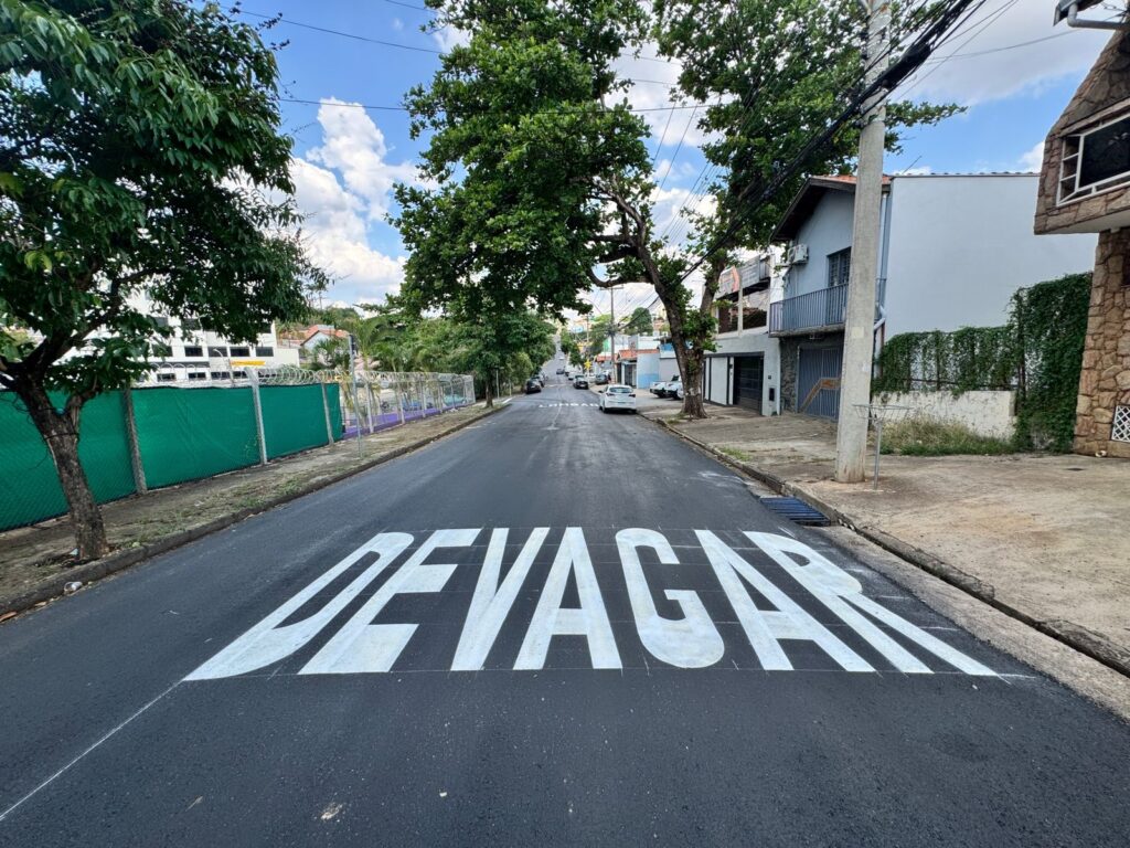 A imagem mostra uma rua residencial com a palavra "DEVAGAR" pintada em letras brancas grandes e em negrito no asfalto. A rua é ladeada por árvores frondosas e casas. Há carros estacionados e em movimento na rua, bem como postes de eletricidade e fios aéreos. O céu está azul com algumas nuvens brancas. A pintura no asfalto sugere que esta é uma área onde os motoristas devem reduzir a velocidade, possivelmente perto de uma escola, parque ou cruzamento movimentado.