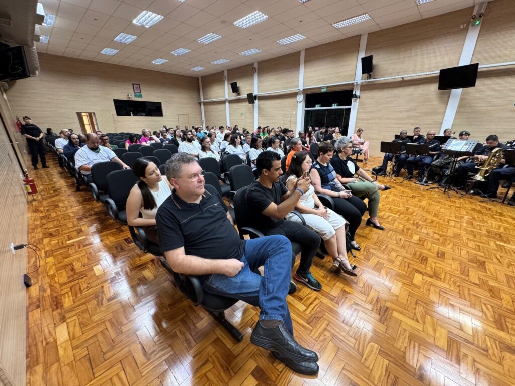 A imagem mostra uma plateia sentada em cadeiras em uma sala com piso de madeira e paredes revestidas de madeira. No fundo, à direita, há uma banda militar tocando instrumentos. A iluminação vem de luminárias no teto. Há algumas pessoas em pé na lateral esquerda da imagem, incluindo um guarda. A atmosfera parece ser de um evento formal ou apresentação.
