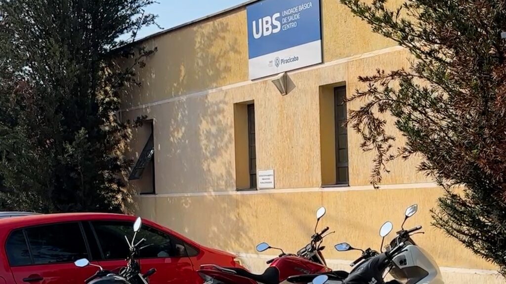 A imagem mostra a fachada de um prédio com um letreiro azul que diz "UBS UNIDADE BÁSICA DE SAÚDE CENTRO PIRACICABA". O prédio é de cor amarela, com algumas janelas e arbustos em frente. Há também um carro vermelho e algumas motocicletas estacionadas. A luz do sol projeta sombras no prédio.