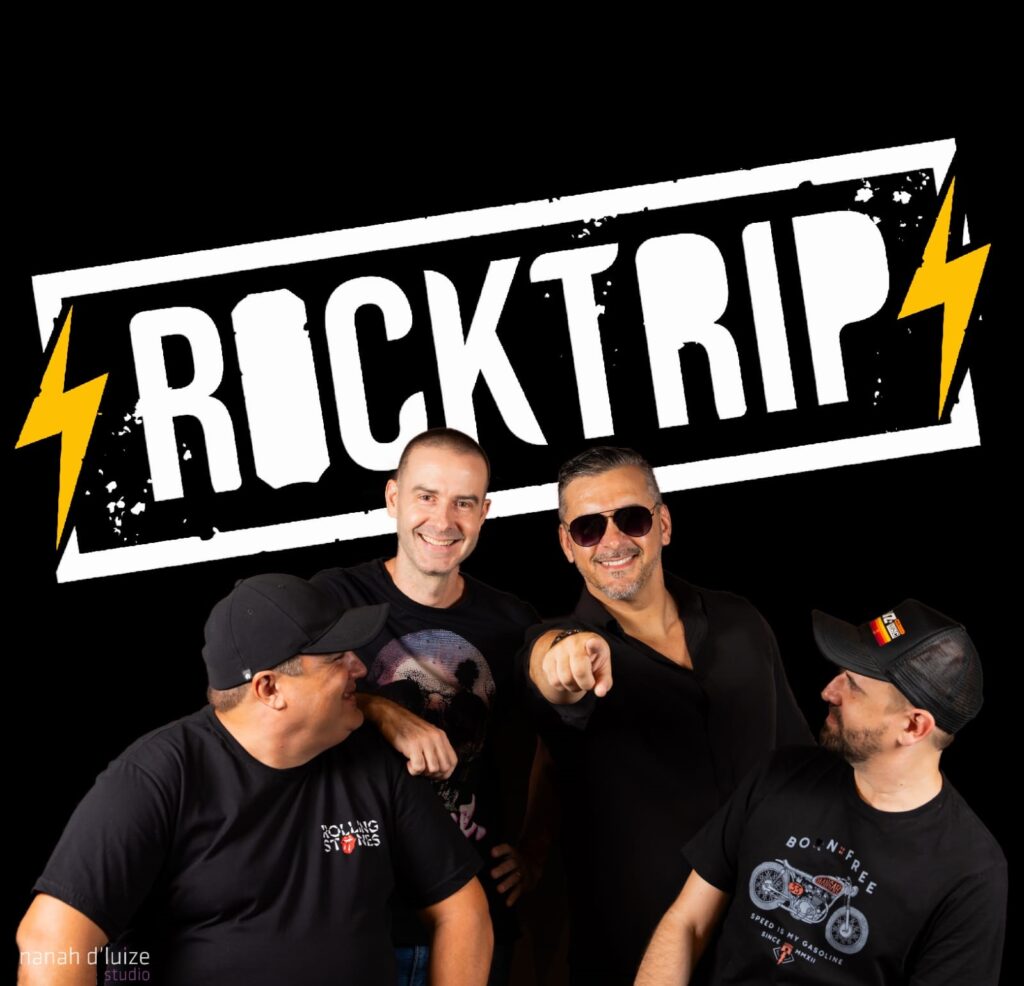 A imagem mostra quatro homens em frente a um fundo preto. Acima deles, há uma faixa branca com as letras "ROCKTRIP" em letras maiúsculas brancas e grossas, com raios amarelos em cada lado. Os homens estão vestidos com camisetas pretas. O homem mais à esquerda usa um boné preto e olha para o homem ao seu lado. Este homem, o segundo da esquerda, também usa uma camiseta preta com uma estampa colorida e sorri para a câmera, com o braço apoiado no ombro do homem à sua esquerda. O terceiro homem da esquerda, em destaque no centro, usa uma camisa preta desabotoada e óculos escuros, apontando com o dedo indicador para a frente e sorrindo. O homem mais à direita usa um boné preto com uma bandeira espanhola e olha para o homem ao seu lado, sorrindo levemente. Sua camiseta exibe uma estampa de uma motocicleta vintage com a frase "BORN FREE" e outros textos.