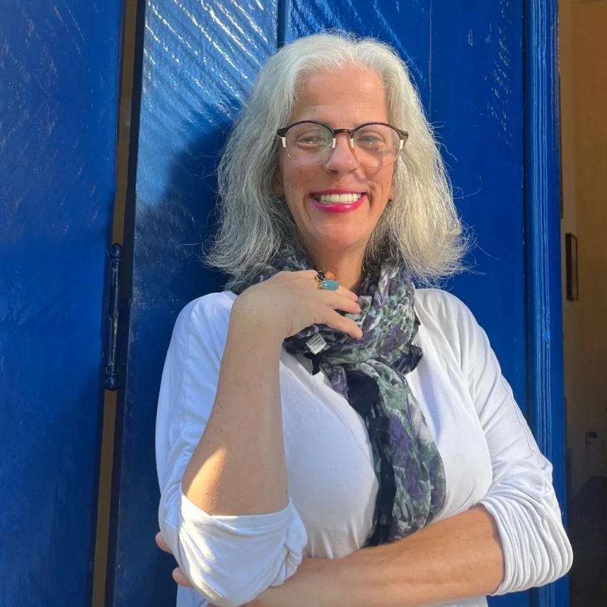 A imagem mostra uma mulher com cabelos grisalhos, óculos e um sorriso radiante, vestindo uma blusa branca e um lenço estampado. Ela está em frente a uma vibrante porta azul, com a mão apoiada no pescoço, exibindo anéis chamativos. A iluminação suave realça seus traços e o fundo colorido cria um contraste interessante.