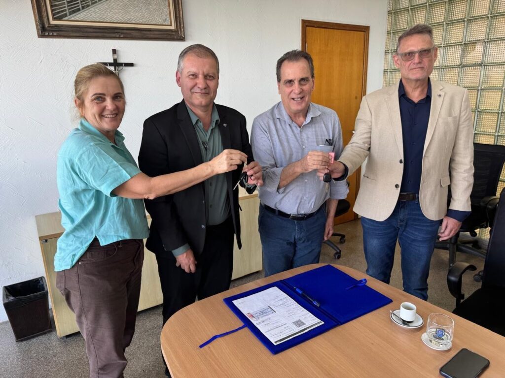 A imagem mostra quatro pessoas em um escritório, em frente a uma mesa de madeira. Duas mulheres e dois homens estão em pé. Um homem de terno preto e camisa verde entrega um molho de chaves a um homem de camisa xadrez cinza e calça jeans. Uma mulher de camisa azul clara e calça marrom sorri para a câmera, enquanto um homem de blazer bege, camisa azul marinho e óculos de sol, também sorri. Na mesa, há uma pasta azul aberta com documentos e uma caneta, uma xícara de café e um celular. Ao fundo, observa-se um quadro com uma imagem, um crucifixo na parede, uma porta de madeira e cadeiras de escritório. A cena parece registrar a entrega de chaves ou um acordo formal.