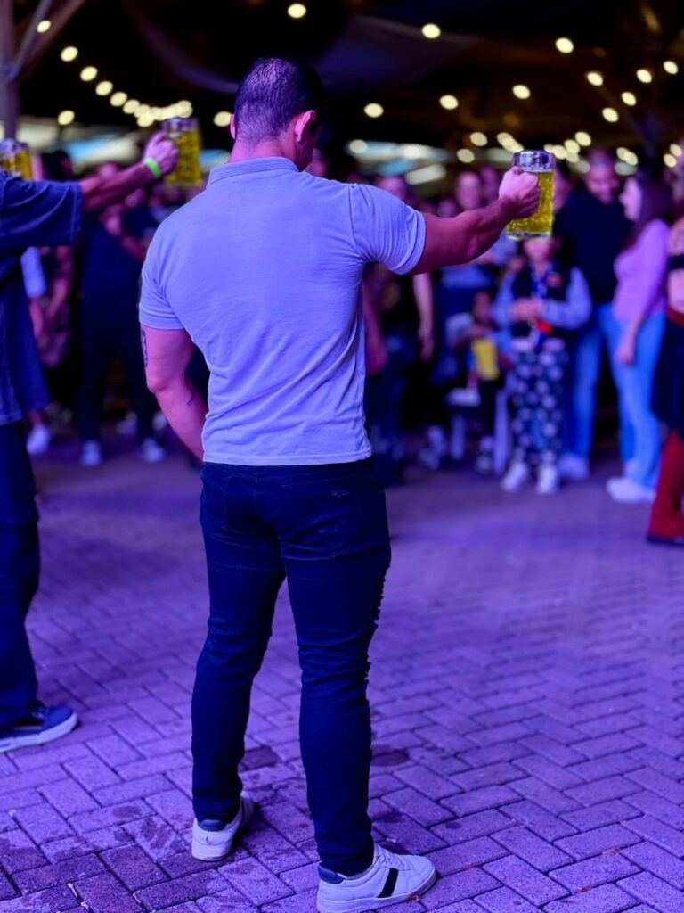 A imagem mostra um homem de costas, segurando um copo de cerveja com a mão direita erguida. Ele está usando uma camisa polo cinza e calças jeans escuras, com tênis brancos. O ambiente parece ser uma festa ou evento ao ar livre, com luzes de fada penduradas no teto e uma multidão de pessoas ao fundo. O chão é pavimentado com tijolos e há uma iluminação roxa predominante.