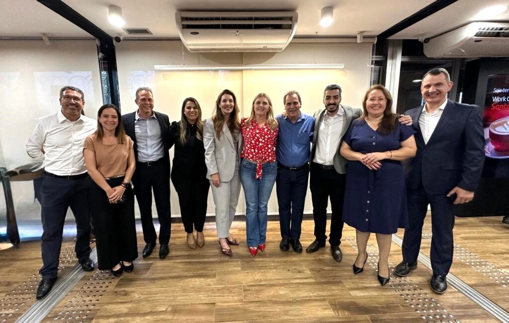 A imagem mostra um grupo de dez pessoas, seis homens e quatro mulheres, posando juntos em um ambiente interno. Todos estão em pé e parecem estar em uma reunião ou evento. Da esquerda para a direita: * O primeiro homem está vestindo uma camisa branca de manga longa, calças azuis escuras e sapatos pretos. Ele usa óculos e tem um sorriso. * A primeira mulher, ao lado dele, veste uma blusa marrom de manga curta, uma saia preta longa e sapatos pretos. Ela tem uma bolsa preta na mão. * O segundo homem está vestindo uma camisa cinza clara de manga longa, calças pretas e sapatos pretos. * A segunda mulher está vestindo um terno cinza claro, composto por blazer e calça, e sapatos com estampa de oncinha. * A terceira mulher está vestindo uma blusa vermelha com estampa de bolinhas brancas, calça jeans azul e sapatos vermelhos. * O terceiro homem está vestindo uma camisa azul de manga longa, calças jeans azuis e sapatos pretos. * O quarto homem está vestindo uma camisa branca de manga longa e uma calça preta. Ele tem um blazer cinza sobre os ombros. * A quarta mulher está vestindo um vestido azul marinho de manga curta, com botões na frente, e sapatos pretos. * O quinto homem, à direita, está vestindo uma camisa branca de manga longa e um terno azul escuro, com calças combinando e sapatos pretos. Ele tem um braço em volta do ombro da mulher ao seu lado. O fundo da imagem mostra um teto com luminárias e um aparelho de ar condicionado. Há também uma tela ao fundo com uma imagem de uma xícara de café e texto em português. O chão é de madeira com um padrão de listras em relevo.