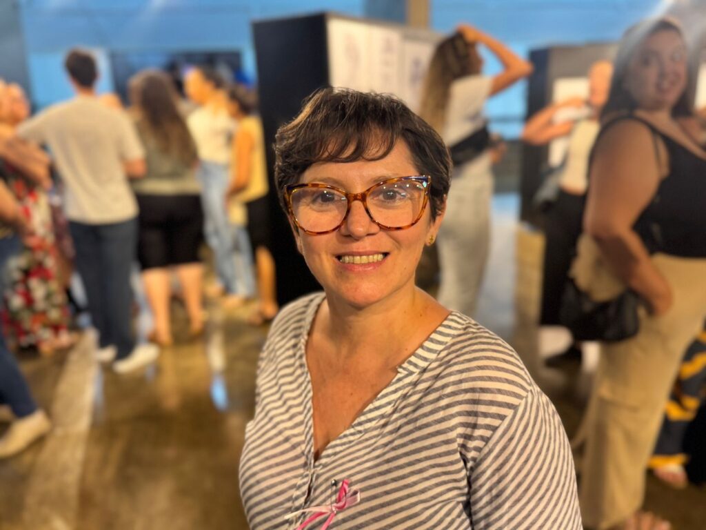 A imagem mostra uma mulher sorridente com óculos de armação tartaruga e cabelo curto e escuro. Ela está usando uma blusa listrada em tons de cinza e branco, com um broche de laço rosa preso na gola. O fundo da imagem está desfocado, mas é possível ver outras pessoas em segundo plano, sugerindo que ela está em um evento ou reunião social. A iluminação parece ser artificial e suave, criando uma atmosfera acolhedora.