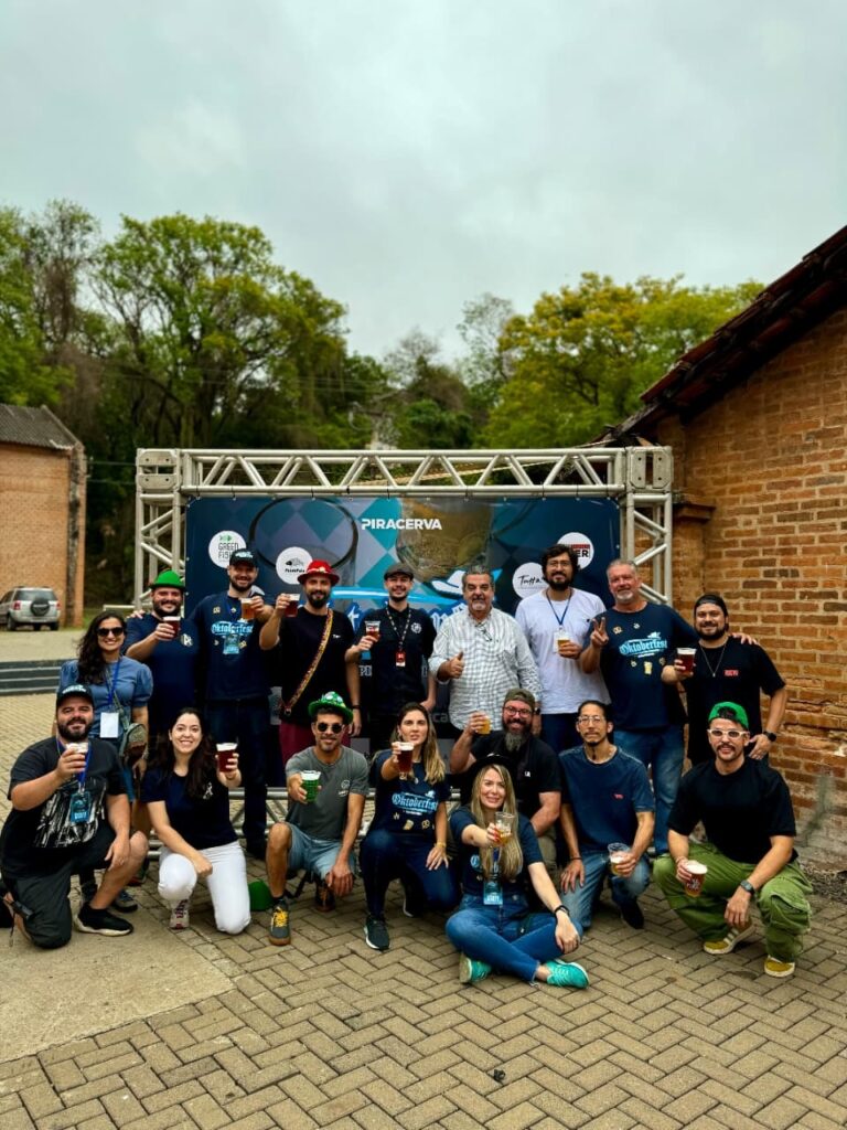 A imagem mostra um grupo de pessoas em frente a um palco, com uma faixa escrita "PIRACERVA". Todos parecem estar celebrando e muitos seguram copos de cerveja. A maioria está vestida casualmente, alguns com chapéus temáticos, sugerindo um evento festivo, possivelmente uma Oktoberfest. O ambiente é ao ar livre, com árvores ao fundo e uma construção de tijolos à direita. As pessoas estão em diferentes posições, algumas em pé e outras agachadas, todas sorrindo e interagindo.