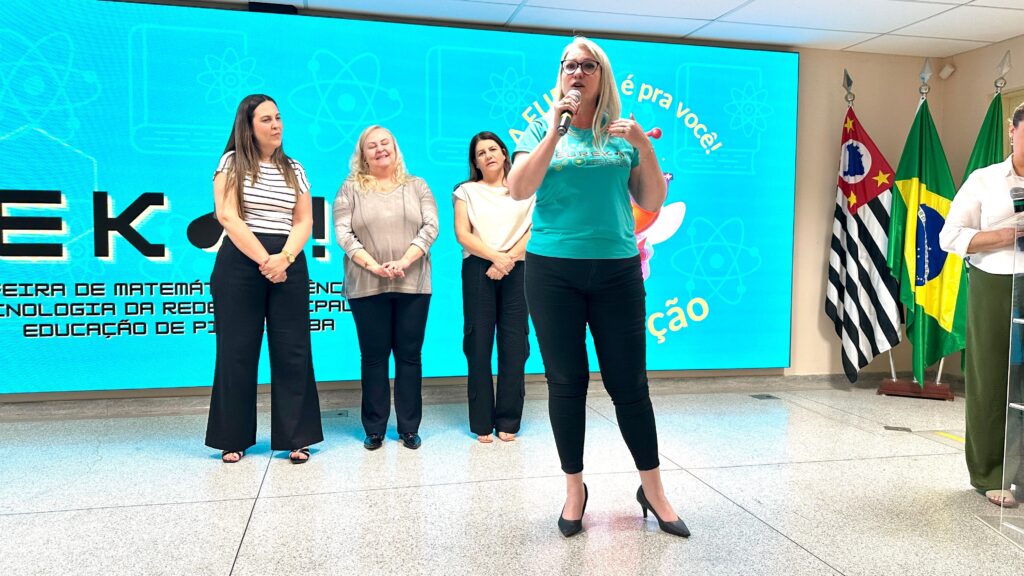 A imagem mostra um grupo de quatro mulheres em pé em frente a um telão azul brilhante. A mulher mais à direita está falando em um microfone, usando uma camiseta verde com a palavra "EUREKA" impressa nela. As outras três mulheres estão em pé ao lado dela, olhando para ela ou para o público. O telão atrás delas exibe o texto "EK!" em letras grandes e brancas, com desenhos de átomos e livros espalhados. Abaixo disso, há texto em português que parece ser o nome de um evento, possivelmente relacionado a feiras de matemática, tecnologia e educação. À direita do telão, há três bandeiras, incluindo a bandeira do Brasil e a bandeira do estado de São Paulo. A iluminação geral da sala é clara, com uma luz suave vindo de cima. O piso é um material liso e brilhante, possivelmente mármore ou granito polido. As mulheres estão vestidas de forma casual, mas profissional. A atmosfera geral parece ser de um evento educacional ou de apresentação.