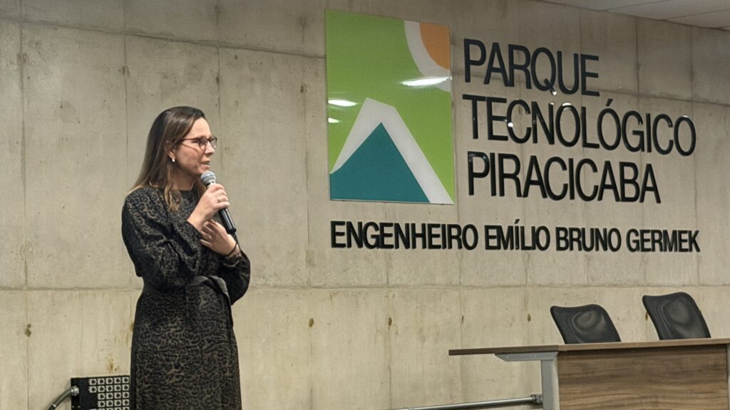 Uma mulher está em pé, falando ao microfone, diante de uma parede de concreto com uma placa que diz “Parque Tecnológico Piracicaba Engenheiro Emílio Bruno Germek”. Ao lado dela há um pódio de madeira com outro microfone, e atrás, várias cadeiras organizadas em fileiras, sugerindo um evento formal como uma palestra ou cerimônia. A placa tem um logotipo colorido com formas em verde, laranja, azul e branco. O ambiente é iluminado e transmite seriedade e profissionalismo.