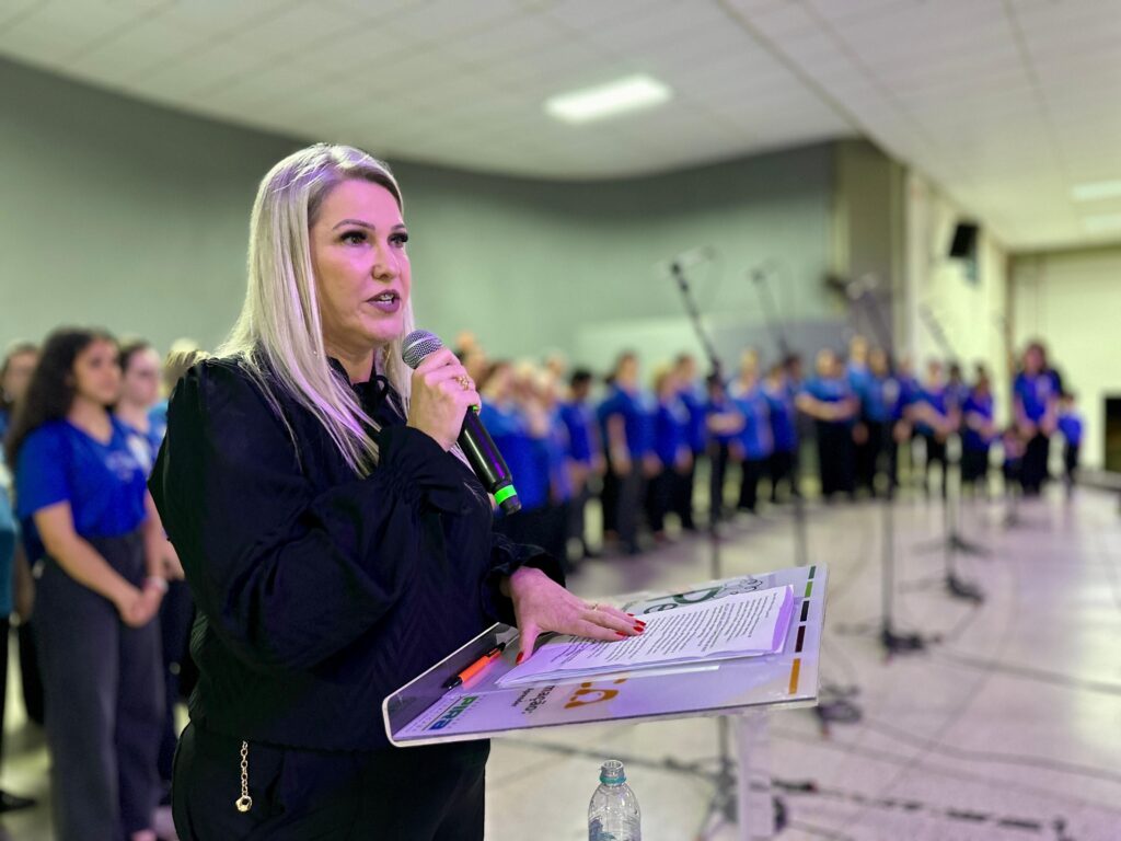 A imagem mostra uma mulher loira com cabelo comprido, usando um microfone e falando para uma plateia, que parece ser um coro de jovens vestindo camisetas azuis. Ela está em primeiro plano, segurando um pedestal com papéis. Seu cabelo é loiro platinado e ela usa maquiagem. Ela está vestindo uma blusa preta de mangas compridas com detalhes bufantes nos ombros e punhos. O coro está ao fundo, em foco desfocado, e parece estar em um palco ou auditório. A iluminação geral é suave, com tons verdes e roxos, sugerindo um ambiente de performance.