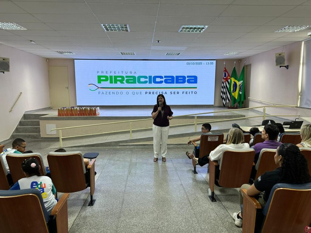 A imagem mostra uma sala de conferências onde uma mulher está apresentando para uma audiência. A mulher, que está em pé no centro da sala, usa uma camisa roxa escura e calças brancas. Ela está segurando um microfone e falando para um grupo de pessoas sentadas em cadeiras de madeira voltadas para ela. No fundo da sala, uma grande tela exibe o logotipo "PREFEITURA PIRACICABA" com o slogan "FAZENDO O QUE PRECISA SER FEITO". Ao lado da tela, há três bandeiras: a bandeira do estado de São Paulo, a bandeira do Brasil e a bandeira do município de Piracicaba. A audiência parece ser composta por um grupo diversificado de pessoas, incluindo jovens e adultos. Alguns dos jovens parecem ser estudantes, como indicado pelas camisetas com o logo "Edu". A iluminação da sala é uniforme, vinda do teto, e a atmosfera geral sugere um evento educacional ou informativo.