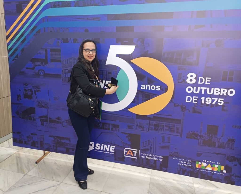 A mulher está em frente a um painel comemorativo. O painel tem um fundo azul com fotos em preto e branco de prédios e pessoas. Há um grande número "5" em branco e verde, com um semicírculo laranja ao lado, representando 50 anos. À direita do "50 anos", está escrito "8 DE OUTUBRO DE 1975" em letras brancas. Abaixo do "50 anos", há logotipos do SINE, FAT e do Governo do Brasil. A mulher, usando óculos, uma blusa preta e calças azuis, segura uma medalha em uma caixa aberta. Ela sorri para a câmera.