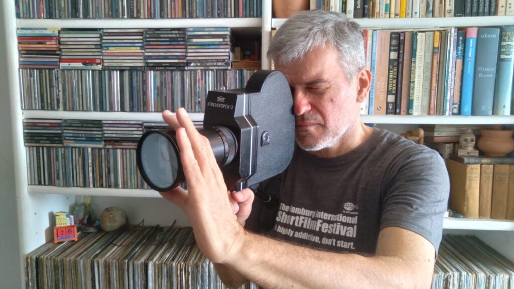 A imagem mostra um homem de cabelos grisalhos olhando através de uma câmera de cinema antiga, um Krasnogorsk-3. Ele está em frente a uma estante cheia de CDs, discos de vinil e livros. A câmera está posicionada para filmar, com a mão esquerda segurando a lente e a mão direita apoiando o corpo da câmera. O homem está usando uma camiseta cinza com uma estampa que diz "The Hamburg International ShortFilmFestival - highly addictive, don't start." O fundo é dominado por prateleiras repletas de mídias físicas. Na prateleira acima das mídias, há uma coleção de livros com títulos em vários idiomas. Na prateleira inferior, entre os discos de vinil, há alguns objetos decorativos, como um pequeno calendário com o número 25, um frasco de vidro, uma pedra e uma pequena escultura de caveira. A iluminação é suave, vindo provavelmente de uma janela lateral, criando um ambiente acolhedor e nostálgico.