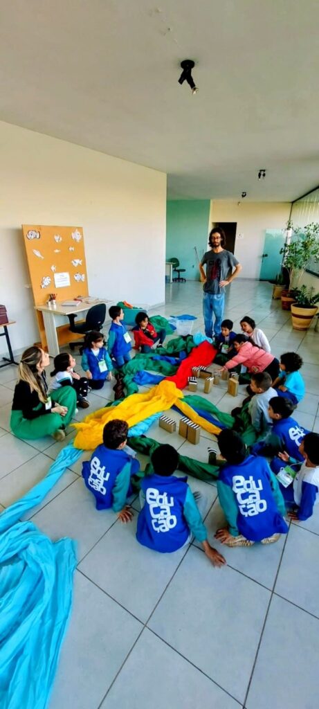 A imagem mostra um grupo de crianças e dois adultos sentados no chão em uma sala. As crianças estão vestidas com uniformes azuis com a palavra "Educação" escrita. Elas estão cercadas por tecidos coloridos, como azul, verde, amarelo e vermelho, e algumas caixas de papelão que parecem ser usadas em uma atividade. Um homem adulto, com barba e cabelo escuro, está em pé no centro da sala, de costas para a câmera, com as mãos nos quadris. Uma mulher adulta, com cabelo loiro, está sentada no chão à esquerda, interagindo com algumas crianças. Ao fundo, há uma mesa com materiais educativos, como livros e desenhos de peixes em um quadro. A sala parece ser um espaço de aprendizado, possivelmente uma sala de aula ou um centro de atividades para crianças. A iluminação é natural, vindo de uma janela ou porta de vidro ao fundo.