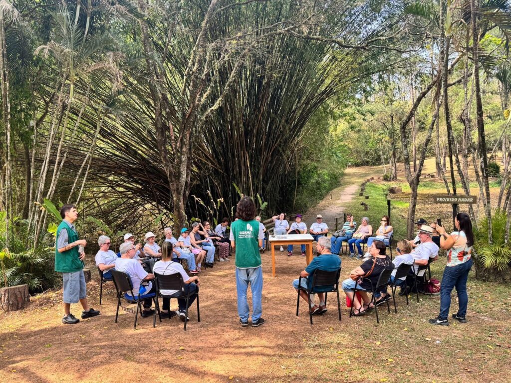 A imagem mostra um grupo de pessoas sentadas em cadeiras ao ar livre, em um ambiente natural com muita vegetação, incluindo bambus altos e árvores. Há duas pessoas em pé, possivelmente guias ou palestrantes, conversando com o grupo. Uma delas, vestida com um colete verde com o logo "SEDEMA", está de costas para a câmera, falando com as pessoas sentadas. A outra pessoa, vestida com um colete verde e bermuda, está de frente para a câmera, também falando com o grupo. O cenário é um caminho de terra que serpenteia pela paisagem verde. Ao fundo, há uma placa com os dizeres "PROIBIDO NADAR", indicando a presença de um corpo d'água próximo. A iluminação sugere que é um dia ensolarado. As pessoas no grupo parecem estar participando de uma palestra, tour guiado ou alguma atividade educativa ao ar livre.