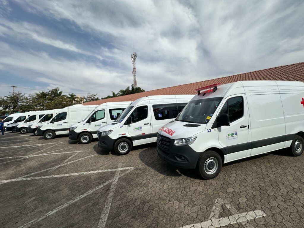 A imagem mostra uma fila de ambulâncias brancas estacionadas em um pátio de concreto com marcações de estacionamento. As ambulâncias são todas do mesmo modelo, parecendo ser vans Mercedes-Benz Sprinter. A ambulância mais próxima da câmera, à direita, tem um grande sinal de "+" vermelho na lateral e a palavra "AMBULÂNCIA" escrita em letras vermelhas na frente. Ela também tem uma luz vermelha intermitente no teto. Há um número "33" pintado na porta do motorista. Um logotipo com as palavras "PREFEITURA" e "PIRA" está visível na lateral. As outras ambulâncias na fila também são brancas e parecem ter o mesmo logotipo. Algumas têm números pintados nas portas, como "17", "26" e "08". A ambulância com o número "26" tem um símbolo de acessibilidade para cadeira de rodas. Ao fundo, há um edifício com telhado de telhas marrons e árvores. Uma torre de telecomunicações com antenas é visível no céu, que está parcialmente nublado com nuvens brancas e cinzas. O sol parece estar brilhando entre as nuvens.