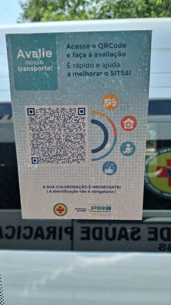 A imagem mostra um adesivo colado em uma janela, provavelmente de um veículo de transporte. O adesivo tem um fundo azul claro e contém texto e um QR code. Na parte superior esquerda, em letras grandes, está escrito "Avalie nosso transporte!". Ao lado, em texto menor, lê-se "Acesse o QRCode e faça a avaliação. É rápido e ajuda a melhorar o SITSS!". No centro da imagem, há um QR code grande que pode ser escaneado para acessar a avaliação. À direita do QR code, há ícones gráficos que parecem representar diferentes aspectos do serviço, como um veículo de emergência, uma casa com uma cruz vermelha (indicando saúde), uma pessoa segurando um bebê e um polegar para cima (indicando aprovação ou satisfação). Na parte inferior do adesivo, há uma mensagem em letras maiúsculas: "A SUA COLABORAÇÃO É IMPORTANTE! | A identificação não é obrigatória |". Abaixo dessa mensagem, há logotipos, incluindo um brasão com uma cruz vermelha e os dizeres "SECRETARIA DE SAÚDE" e "PREFEITURA PIRA RACICABA". A imagem também reflete o ambiente externo através da janela, com algumas folhas verdes visíveis no topo e o reflexo de um letreiro com texto em português na parte inferior.