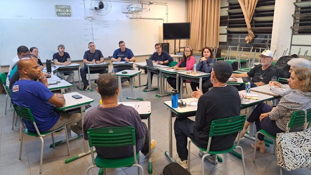 A imagem mostra uma sala de aula com várias pessoas sentadas em carteiras, participando de uma reunião ou aula. A iluminação parece ser artificial, vinda de luminárias no teto, e há luz natural entrando pelas janelas ao fundo. No quadro branco, há algumas anotações e um pequeno cartaz com a inscrição "GENTILEZA + GERA GENTILEZA". Há também um ventilador de teto e um projetor. As pessoas sentadas nas carteiras são de diferentes idades e gêneros, e algumas parecem estar engajadas em discussões, enquanto outras estão ouvindo atentamente. Várias carteiras possuem cadernos, canetas e alguns objetos pessoais, como garrafas de água e celulares. Ao fundo, há uma estante com cadeiras empilhadas e cortinas nas janelas. A atmosfera geral sugere um ambiente de aprendizado ou colaboração.