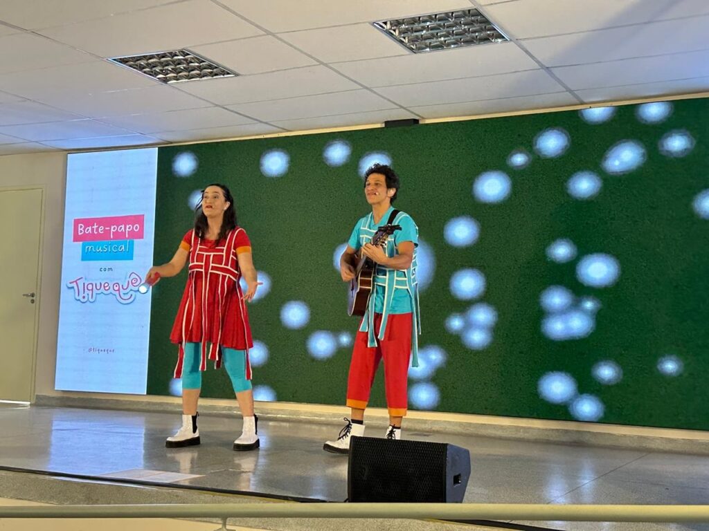 A imagem mostra um palco com dois artistas se apresentando. À esquerda, uma mulher com um vestido vermelho vibrante e leggings azuis claras gesticula enquanto fala. Ao lado dela, um homem toca violão, vestindo uma camisa azul turquesa com detalhes brancos e calças vermelhas. Um telão à esquerda exibe o texto "Bate-papo musical com Tiqueque", com o logotipo do Tiqueque em destaque. O fundo do palco é um painel verde com projeções de círculos brancos luminosos, lembrando bolhas ou luzes. Ambos os artistas usam sapatos chamativos, combinando com suas roupas coloridas. Ao primeiro plano, um alto-falante preto está posicionado. A iluminação sugere que a apresentação está ocorrendo em um ambiente interno.