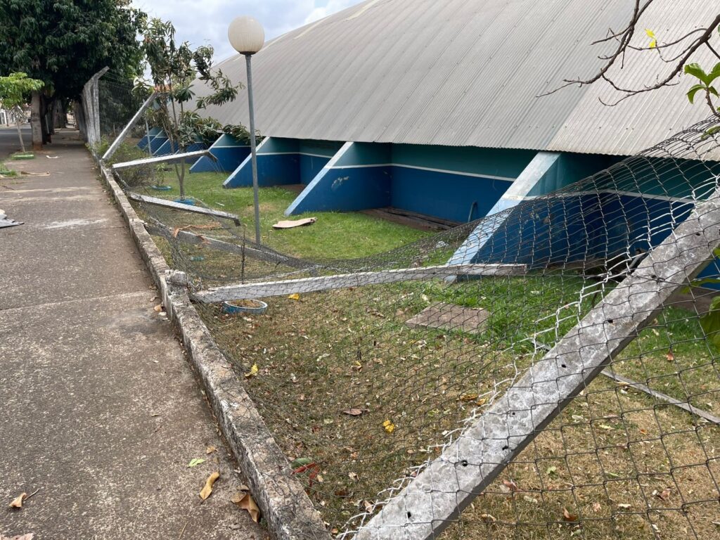 A imagem mostra uma cena externa com uma calçada de concreto à esquerda e um gramado à direita, que faz parte de um terreno adjacente a uma edificação. Uma cerca de arame, que parece ter sido danificada, atravessa a imagem em primeiro plano, com postes de concreto inclinados e o arame solto em alguns pontos. A edificação ao fundo possui um telhado de metal ondulado e uma estrutura com janelas ou aberturas em arco, pintada em tons de azul e verde. Há uma luminária de poste com um globo branco em frente à edificação. Algumas árvores e galhos de árvores também são visíveis, assim como folhas caídas no gramado e na calçada. O céu está nublado.