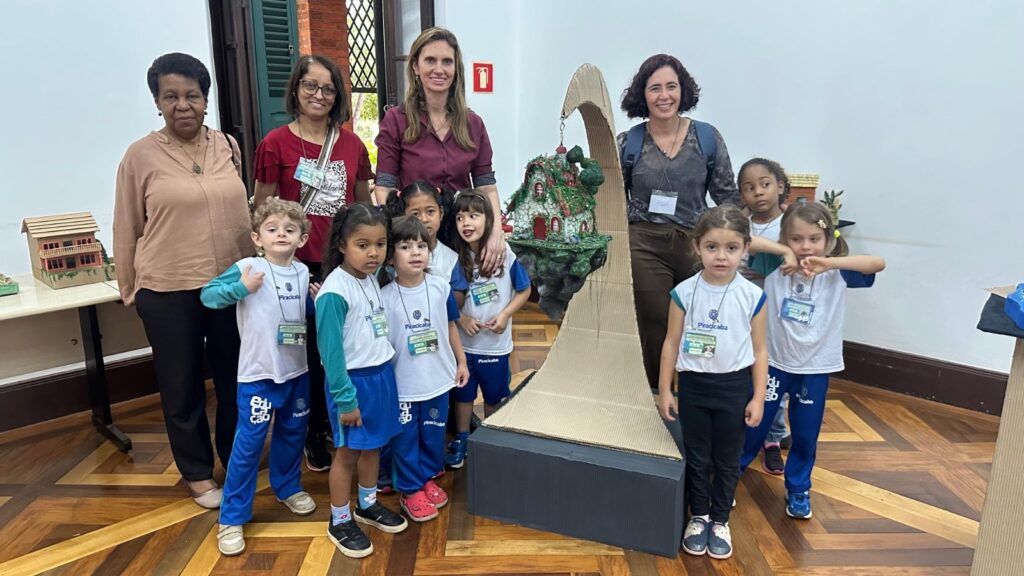 A imagem mostra um grupo de mulheres adultas e crianças em pé em frente a uma construção feita de papelão. As mulheres estão atrás das crianças, e a maioria das crianças está usando uniformes escolares com crachás. Há uma casa em miniatura em uma mesa à esquerda da imagem, e a construção de papelão parece ser uma espécie de rampa ou arco com uma casa em miniatura no topo. O piso é de madeira com um padrão geométrico.