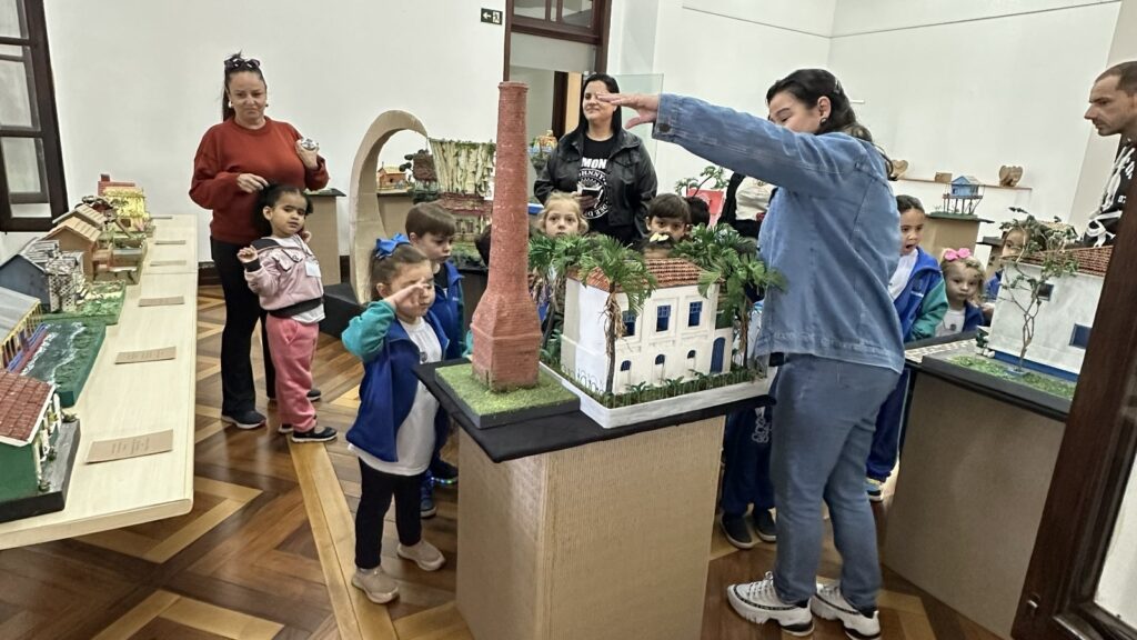 A imagem mostra um grupo de crianças e adultos em uma exposição, possivelmente de maquetes. No centro da imagem, uma mulher de jaqueta jeans está explicando um modelo de casa com um chaminé em destaque. As crianças, algumas com uniformes escolares, observam atentamente, demonstrando interesse.À esquerda, uma mulher com um suéter vermelho e uma menina ao seu lado observam uma série de maquetes menores dispostas em uma mesa comprida. As maquetes parecem representar diferentes construções e cenários, com detalhes que sugerem um contexto histórico ou arquitetônico.O ambiente parece ser um museu ou centro cultural, com chão de madeira trabalhado e paredes claras. A iluminação é suave, realçando os detalhes das maquetes e as expressões das pessoas. A atmosfera geral é de aprendizado e exploração.