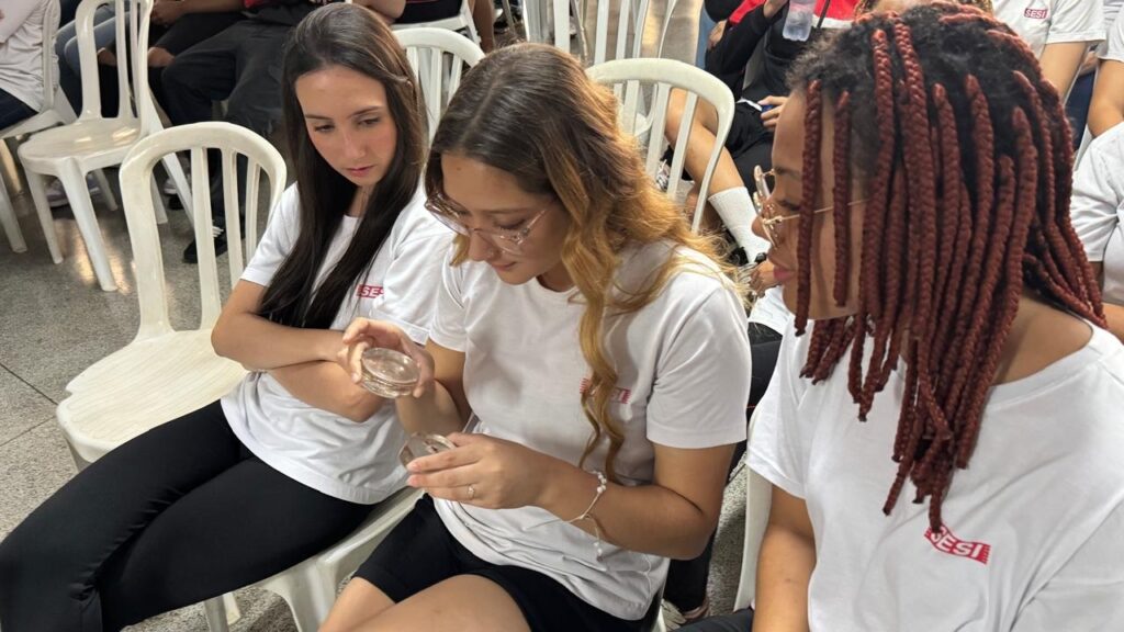 A imagem mostra três jovens sentadas em cadeiras brancas, vestindo camisetas brancas com o logo "SESI" em vermelho. Elas parecem estar em um evento ou atividade, possivelmente em um ambiente educacional ou científico. A jovem da esquerda, com cabelos escuros e compridos, olha para baixo, segurando um pequeno recipiente transparente em suas mãos. A jovem do meio, com cabelos ondulados em tons de castanho e loiro, usa óculos e também está focada em um recipiente transparente que segura. A jovem da direita, com tranças longas em tom avermelhado e óculos, está virada ligeiramente para a esquerda, olhando para as outras duas. O ambiente parece ser interno, com outras pessoas sentadas ao fundo, também em cadeiras brancas. A iluminação é clara, sugerindo um dia ensolarado ou um ambiente bem iluminado. A atmosfera geral é de concentração e interesse no que estão observando nos recipientes.