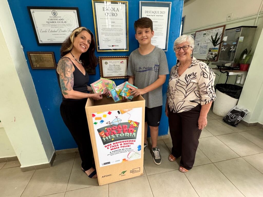 A imagem mostra três pessoas em frente a uma caixa de doações em uma escola. Uma mulher com cabelos escuros e tatuagens nos braços está sorrindo e segurando alguns livros. Ao lado dela, um menino com camiseta cinza e um sorriso tímido observa a caixa. Do outro lado, uma mulher mais velha com óculos e uma blusa estampada também sorri. A caixa de doações tem um cartaz colorido com a mensagem "DOE BRINQUEDOS E FAÇA O NATAL MAIS FELIZ!". Há também certificados e quadros pendurados na parede ao fundo, indicando um ambiente escolar. Ao lado da caixa, há um bebedouro e um saco de lixo. A iluminação sugere que a foto foi tirada em um corredor ou área interna da escola.