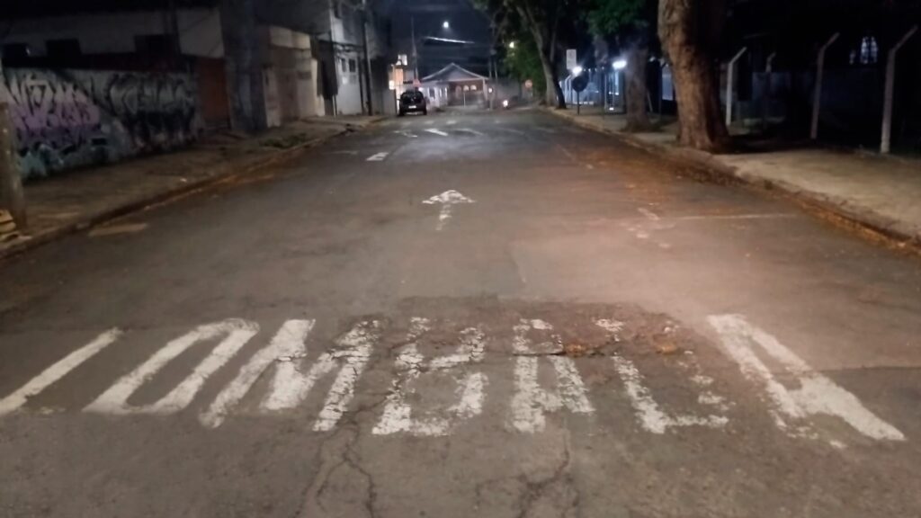 A imagem mostra uma rua à noite, com iluminação pública fraca. No asfalto, há uma faixa branca pintada com a palavra "LOMBADA". A palavra está escrita em letras grandes e desbotadas, indicando que se trata de uma sinalização de trânsito, possivelmente para alertar sobre um quebra-mola ou lombada. O cenário ao redor sugere uma área urbana, com prédios baixos e árvores nas calçadas. Há grafites em um muro à esquerda e um carro estacionado ao longe. A atmosfera geral é de tranquilidade noturna, com a rua aparentemente pouco movimentada.