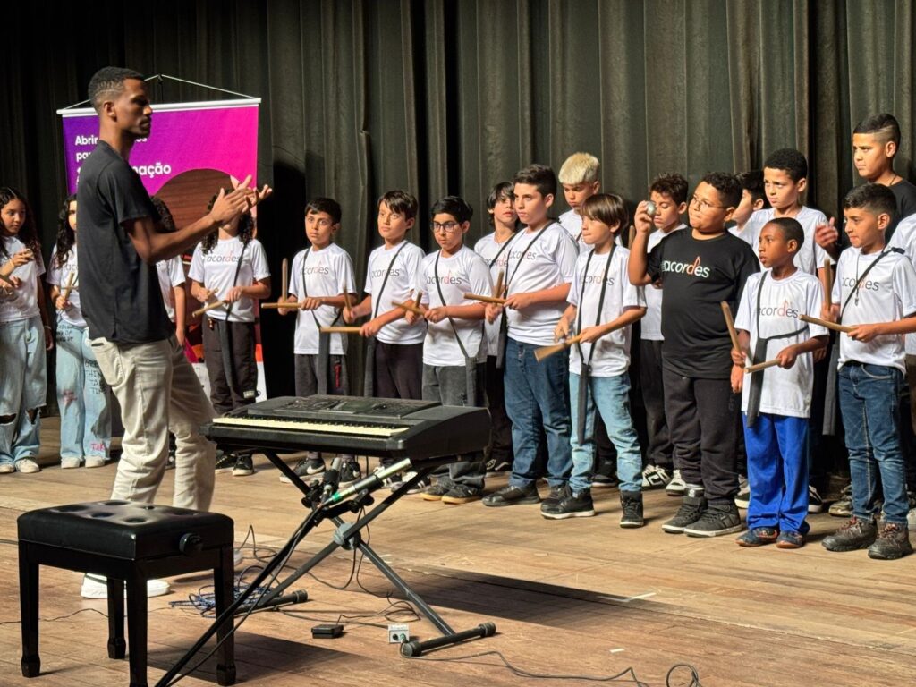 A imagem mostra um grupo de crianças e um adulto em um palco, aparentemente durante uma apresentação musical. O adulto, um homem com pele escura e cabelo curto, está de pé no lado esquerdo do palco, de costas para a câmera, gesticulando com as mãos. Ele veste uma camiseta preta e calças bege claras. À sua frente, um grupo de crianças, a maioria vestindo camisetas brancas com a palavra "acordes" estampada, estão posicionadas em semicírculo. Elas parecem estar participando de uma performance musical, pois várias delas seguram baquetas, sugerindo que estão prestes a tocar algum instrumento de percussão. Um teclado eletrônico está posicionado no centro do palco, em frente às crianças, indicando a presença de acompanhamento musical. O fundo do palco é composto por cortinas escuras, e um banner roxo e rosa com texto em português ("Abrir para ação", com a palavra "ação" parcialmente visível) está pendurado no lado esquerdo, atrás do adulto. A iluminação do palco destaca o grupo, criando uma atmosfera de apresentação. As crianças variam em idade, mas a maioria parece estar na faixa etária escolar. Algumas crianças estão mais à frente, enquanto outras estão mais atrás, formando várias fileiras. A expressão facial de algumas crianças sugere concentração e entusiasmo.