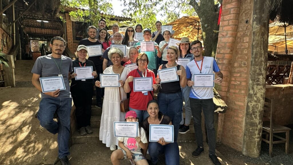 A imagem mostra um grupo de pessoas sorrindo e segurando certificados. Eles estão ao ar livre, em um ambiente com vegetação e construções rústicas de tijolos e madeira. A maioria das pessoas está em pé, em diferentes fileiras, enquanto duas pessoas estão sentadas na frente. Todos parecem felizes e orgulhosos por receberem seus certificados. A luz do sol ilumina a cena, criando um ambiente acolhedor e festivo.