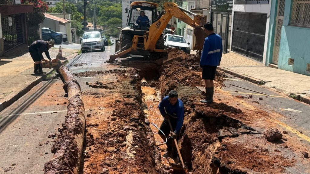 A imagem mostra uma cena de trabalho em uma rua, onde uma grande vala foi escavada no asfalto. Vários trabalhadores estão envolvidos na obra. Em primeiro plano, um trabalhador com um macacão azul está ajoelhado na vala, utilizando uma pá para mexer na terra úmida e avermelhada. Ao lado dele, um cano grande e sujo de terra está parcialmente exposto. Mais atrás, um trator amarelo da marca JCB está posicionado sobre a vala, com seu braço mecânico levantado, indicando que está sendo usado para remover terra ou movimentar materiais. Um trabalhador está no assento do trator, operando a máquina. Na calçada, um outro trabalhador, também vestindo um macacão azul, está em pé e parece estar supervisionando ou aguardando instruções. Ele está usando botas de borracha. Um cano grande e sujo de terra está sendo manuseado por um trabalhador na calçada, que utiliza uma ferramenta para cortá-lo ou ajustá-lo. Ao fundo, carros estão estacionados e passando pela rua, que parece ter um declive acentuado. Edifícios e vegetação compõem o cenário urbano ao redor da obra. O dia está ensolarado.