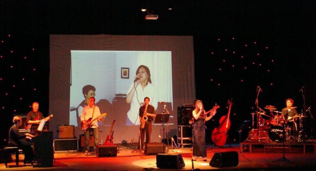 A imagem mostra uma banda se apresentando em um palco. No centro, um telão exibe a imagem de uma mulher cantando, enquanto no palco, uma cantora com um vestido longo e brilhante também canta. Ao lado dela, um músico toca um contrabaixo acústico. Há também um saxofonista, um guitarrista e um pianista no palco. Mais à direita, um baterista está tocando. O fundo do palco é escuro e decorado com pequenas luzes vermelhas. O palco está iluminado com luzes vermelhas e amarelas, criando uma atmosfera de show.