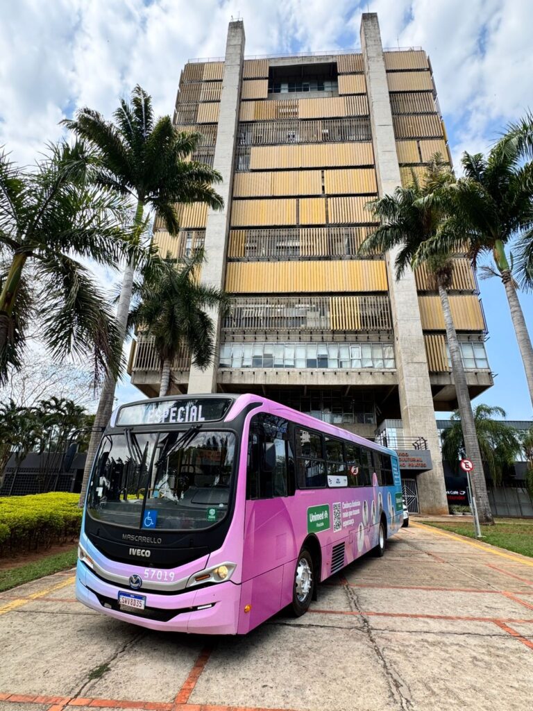 A imagem mostra um ônibus rosa vibrante com detalhes em azul e gradiente, estacionado em frente a um edifício moderno com arquitetura marcante. O ônibus ostenta a palavra "ESPECIAL" em destaque na parte superior e diversas ilustrações ao longo de sua lateral, incluindo o logo da Unimed e um QR code. A lateral do ônibus também exibe o slogan "Cultura histórica que traz nova esperança". O edifício ao fundo é alto e imponente, com uma fachada que combina concreto e elementos metálicos dourados em formato de persianas verticais. Palmeiras altas e exuberantes emolduram a cena, adicionando um toque tropical ao ambiente. O céu está parcialmente nublado, sugerindo um dia claro. A perspectiva da foto, tirada de um ângulo ligeiramente baixo, realça a grandiosidade do edifício e a presença do ônibus. A entrada do edifício é visível, com uma placa indicando "ADO CULTURAL AO PIRATES". Há também um sinal de trânsito vermelho com um 'E' dentro, indicando estacionamento permitido em algumas condições.