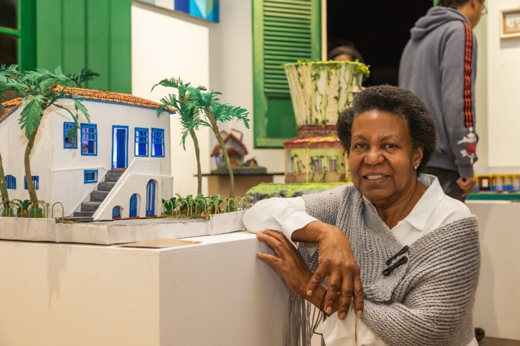 A mulher sorri para a câmera, com o corpo inclinado para a direita, apoiando-se em uma superfície branca. Ela usa uma blusa branca e um xale cinza tricotado sobre os ombros. Ao seu lado esquerdo, há uma miniatura de uma casa branca com telhado laranja e janelas azuis, cercada por palmeiras. Ao fundo, pode-se ver outra miniatura de um edifício, com detalhes em verde e amarelo, e pessoas em segundo plano. A iluminação suave destaca a mulher e as miniaturas, criando uma atmosfera acolhedora.