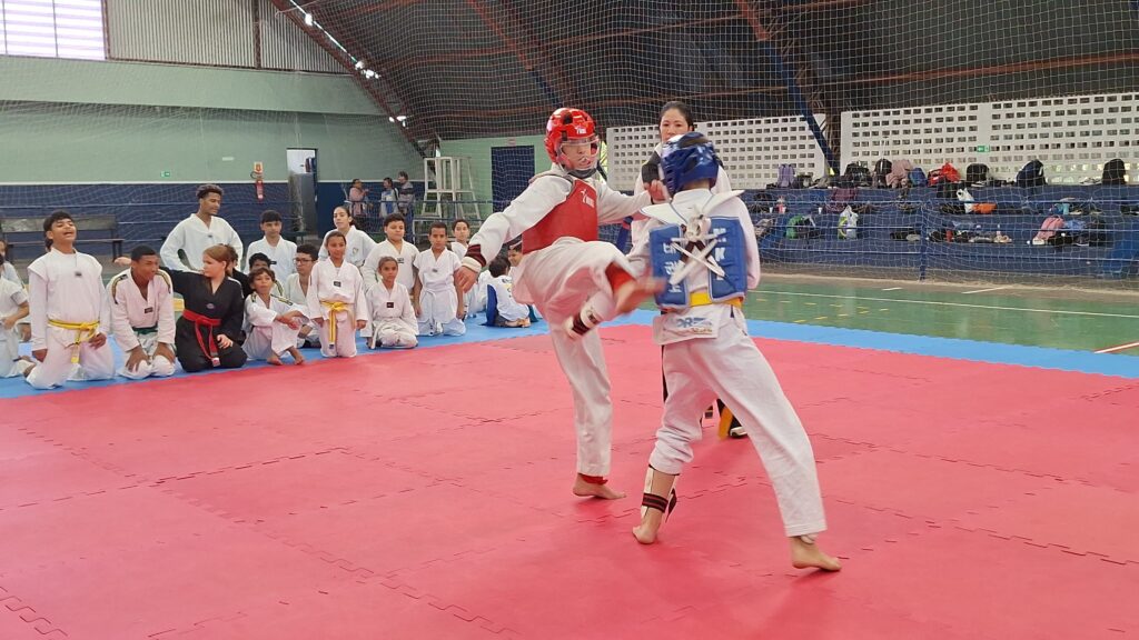 No centro da imagem, dois jovens lutadores estão em ação. Um deles, vestindo um protetor de cabeça vermelho e um colete branco com detalhes vermelhos, está em pleno chute alto com a perna direita. O outro lutador, com um protetor de cabeça azul e um colete azul com detalhes brancos, parece estar se defendendo ou bloqueando o ataque. Ambos vestem o tradicional dobok branco de taekwondo e estão descalços, como é comum na modalidade. Ao redor da área de luta, há outros participantes e espectadores sentados ou ajoelhados. Alguns jovens vestem doboks brancos com faixas coloridas, indicando seus diferentes níveis de graduação. Há também adultos, possivelmente treinadores ou árbitros, observando a luta. Ao fundo, pode-se ver arquibancadas com bolsas e pertences, e a estrutura da quadra com uma rede de proteção. A quadra possui um piso emborrachado nas cores vermelho e azul.
