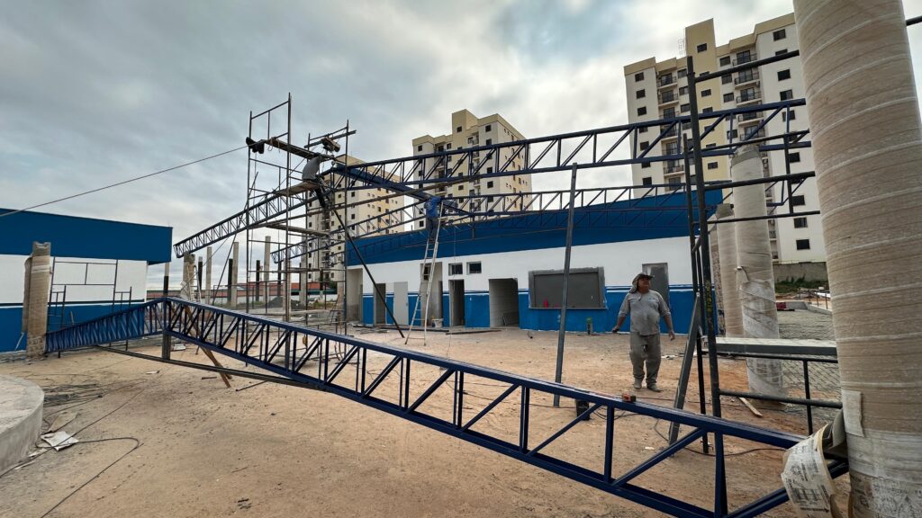 A imagem mostra uma cena de construção em andamento. No primeiro plano, uma estrutura metálica azul, que parece ser parte de um telhado ou marquise, está deitada no chão de terra. Mais atrás, um edifício de um andar com pintura azul e branca está sendo construído ou reformado, com trabalhadores em andaimes instalando a estrutura metálica do telhado. Ao fundo, prédios residenciais altos com vários andares e janelas escuras se destacam contra o céu nublado. Há também pilares cilíndricos cobertos com material de proteção, possivelmente para evitar danos durante a construção. Um trabalhador com chapéu está de pé perto de um dos pilares, observando a obra. A atmosfera geral é de um canteiro de obras ativo.