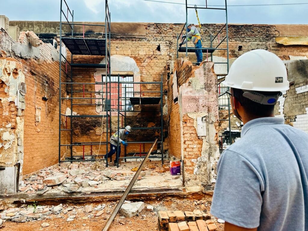 A imagem mostra uma cena de construção em andamento, com uma estrutura de andaimes de metal montada em frente a uma parede de tijolos em ruínas. Várias pessoas estão trabalhando na estrutura. No primeiro plano, à direita, um homem com um capacete branco e uma camisa cinza está de costas, observando a cena. No meio da imagem, há um trabalhador em uma plataforma do andaime, usando um capacete branco, luvas azuis e um colete de alta visibilidade sobre uma camisa cinza. Ele parece estar manuseando algum equipamento ou material. Mais acima no andaime, outro trabalhador, também de capacete e colete, está mais focado na parte superior da estrutura. O chão está coberto de escombros de tijolos e terra, indicando que a construção envolve demolição ou reconstrução. Há também algumas latas de tinta e materiais de construção espalhados pelo local. O céu ao fundo está nublado.