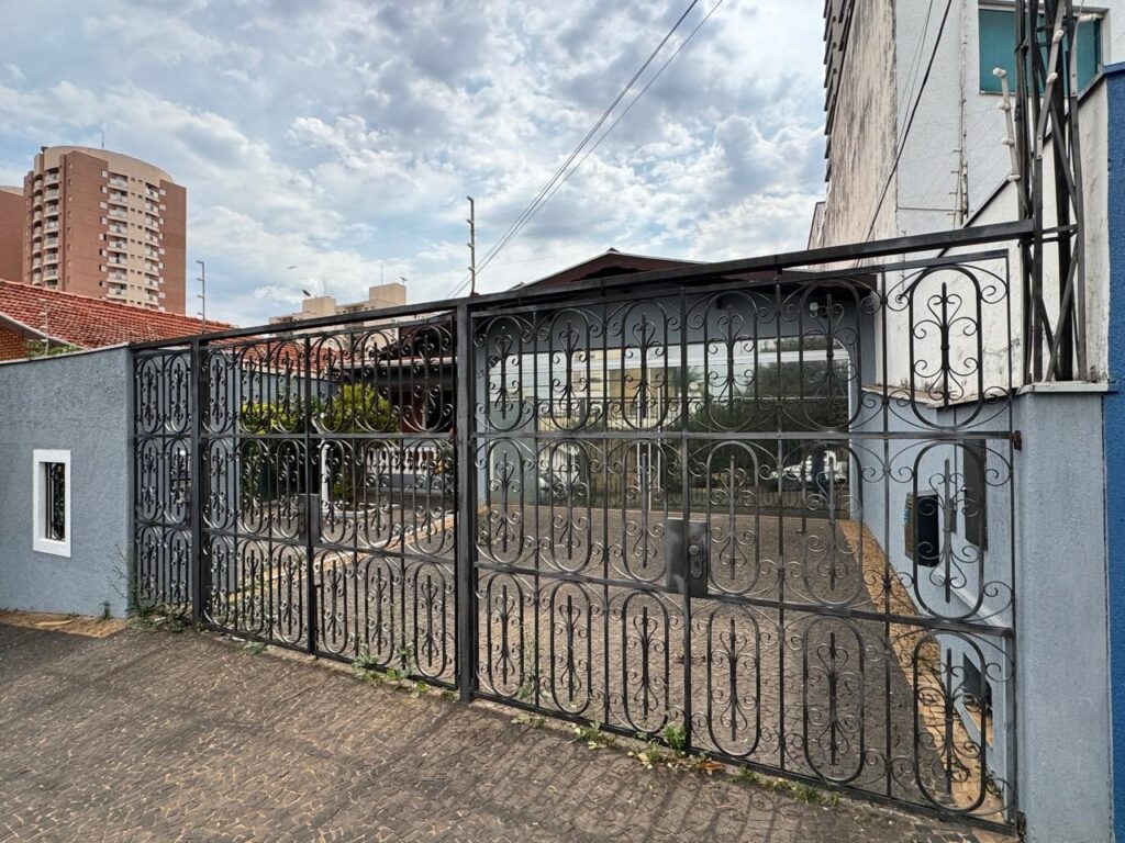 A imagem mostra uma fachada residencial com um muro azul e um portão de ferro forjado. - No muro, há duas janelas retangulares com molduras brancas. - Atrás do muro, um telhado de duas águas cobre a casa. - Um imponente prédio residencial de muitos andares se ergue ao fundo, contrastando com a casa mais baixa. - Fios elétricos atravessam a parte superior da imagem, em meio a um céu com nuvens. - A vegetação, como árvores e arbustos, está presente em alguns pontos da cena.