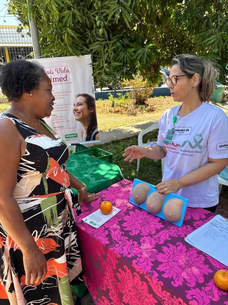 A imagem retrata um evento de saúde ao ar livre, possivelmente uma campanha de conscientização sobre prevenção de câncer de mama, dado o laço rosa visível na camiseta da mulher à direita e o panfleto com informações. Uma mulher afrodescendente, vestindo um vestido estampado em tons de preto, laranja, branco e verde, conversa com uma profissional de saúde. A profissional, usando óculos e uma camiseta branca com o logo da Unimed, aponta para um modelo anatômico de mamas, que está sobre uma mesa coberta por uma toalha rosa com um padrão floral em relevo. A mesa também exibe um pequeno laranja (possivelmente uma tangerina ou laranja kinkan) e panfletos informativos. Ao fundo, uma árvore frondosa com mangas penduradas e um banner com informações sobre "Mais Vida Unimed" reforçam o contexto de saúde e bem-estar. A atmosfera geral é de informação e cuidado.