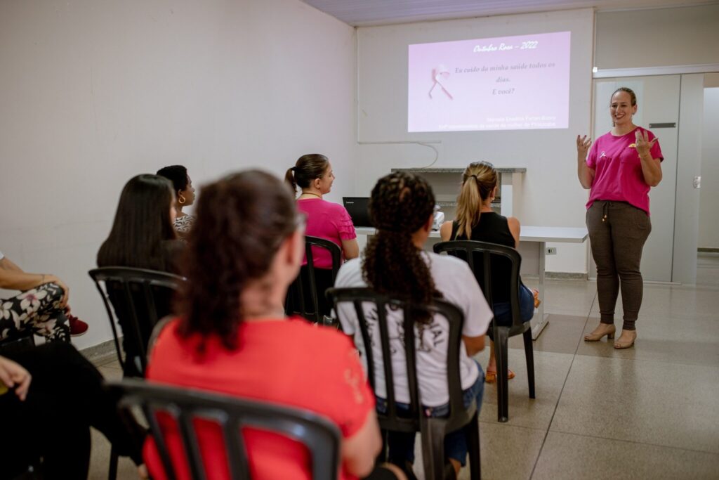 A imagem mostra uma sala de aula ou auditório onde uma mulher está dando uma palestra para um grupo de mulheres. A palestrante, vestindo uma camiseta rosa com um laço rosa no peito, está de pé em frente a uma tela projetada que exibe uma mensagem em português sobre "Outubro Rosa - 2022" e a importância de cuidar da saúde. As mulheres na audiência estão sentadas em cadeiras, voltadas para a tela e para a palestrante. A iluminação é artificial e a sala parece ser um ambiente interno simples, com paredes brancas e piso de concreto. A atmosfera geral é de um evento educativo ou informativo, possivelmente relacionado à conscientização sobre o câncer de mama.