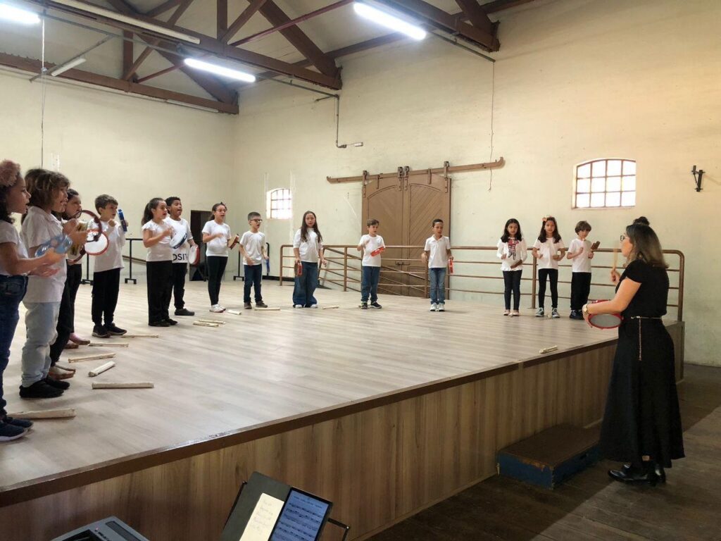 A imagem mostra um grupo de crianças em um palco, participando de uma aula de música. Elas estão em pé, alinhadas, e algumas seguram instrumentos musicais como pandeiros e tambores. Uma professora, de pé à direita, parece estar conduzindo a aula, segurando um tambor e uma baqueta. O palco é de madeira, e ao fundo, há uma grande porta dupla e janelas com treliças. Há também um suporte de partitura no primeiro plano, sugerindo que a aula envolve leitura musical. As crianças vestem roupas casuais, a maioria com camisetas brancas. A atmosfera parece ser de aprendizado e participação ativa.
