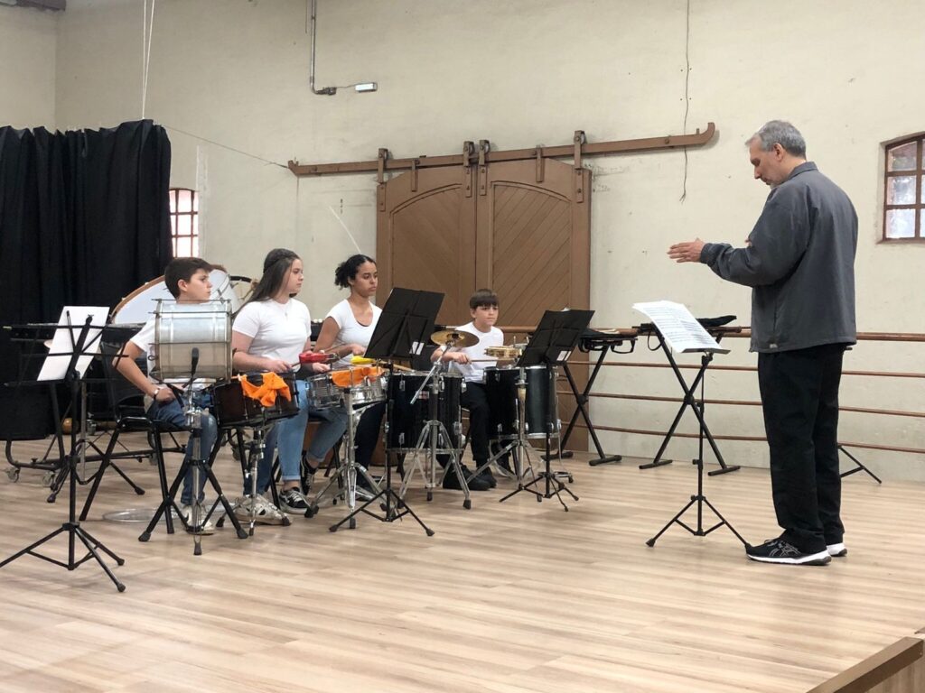 A imagem mostra um grupo de jovens percussionistas em uma sala de ensaio, sob a orientação de um maestro. No primeiro plano, à esquerda, um menino toca um tambor grande. Ao seu lado, uma menina com cabelos longos e castanhos, vestindo uma camiseta branca, toca um tambor menor. Mais à direita, outra menina, com pele morena e cabelos escuros, também vestindo uma camiseta branca, está sentada e parece estar tocando um instrumento de percussão. Um menino mais novo, com uma camiseta branca, está sentado atrás de um conjunto de tambores e pratos, olhando atentamente para o maestro. O maestro, um homem mais velho com cabelos grisalhos e vestindo uma jaqueta cinza e calças pretas, está de pé no lado direito da imagem, de frente para os alunos. Ele gesticula com as mãos, aparentemente dando instruções ou conduzindo a música. Vários estantes de partitura estão posicionados entre os alunos e o maestro.