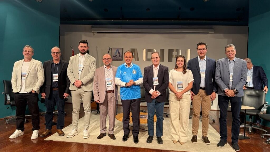 A imagem mostra um grupo de dez pessoas, composto por cinco homens e cinco mulheres, posando para uma foto em um evento. Todos estão vestindo roupas formais ou semi-formais, com crachás visíveis em seus pescoços, indicando que participam de uma conferência ou reunião. Ao fundo, há um logo da "ACIPI". A iluminação é suave e o ambiente parece ser um auditório ou sala de eventos.
